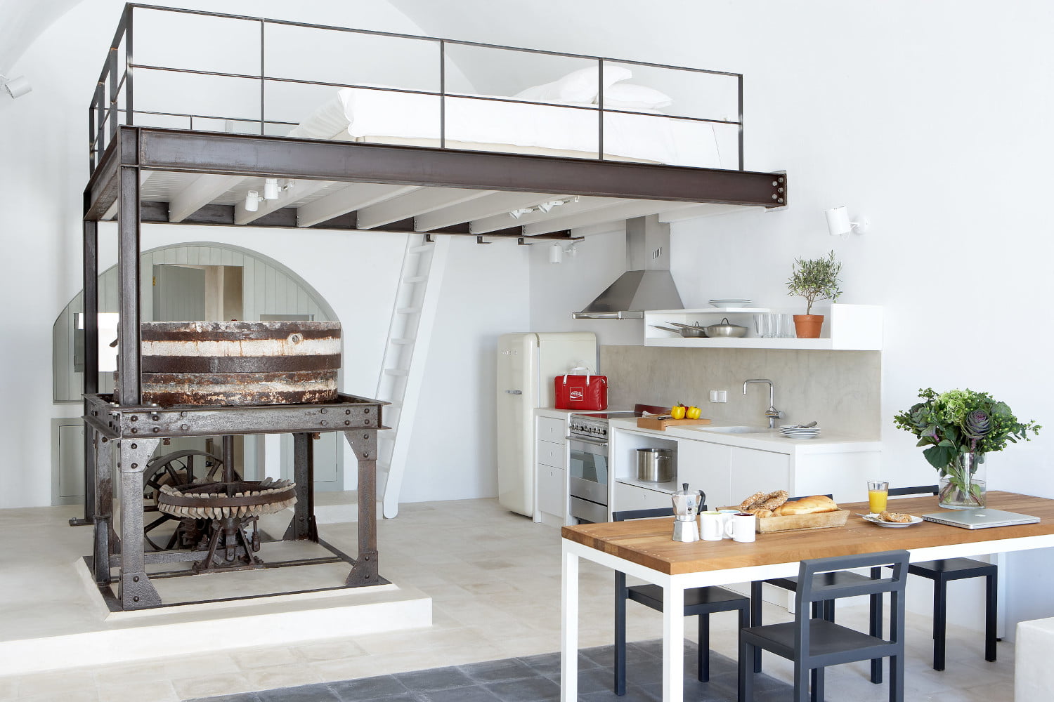 Open plan living area of Villa Mela in Pyrgos Santorini. An old yet preserved piece of machinery from the original factory is on display by the kitchen and dining area, just above the loft were the seconf bedroom is located.