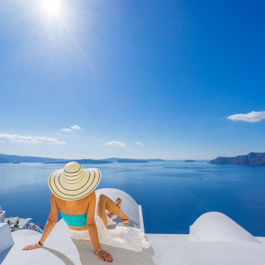 A girl sitting wearing a blue top and a hat sitting on the roof of a Cycladic building in Santorini, overlooking the Aegean Sea and the active volcanos found within the Caldera.
