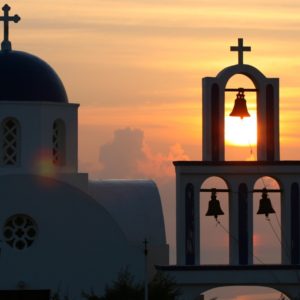 11. A-righteous-cause-Free-Santorini-from-its-sunset-fame-Rent-A-Greek-Villa Photogrpah of a church and its bell tower during sunset in Sabtorini with clouds in the background by Rent A Greek Villa