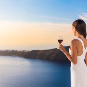 5. A-righteous-cause-Free-Santorini-from-its-sunset-fame-Rent-A-Greek-Villa A girl wearing a white dress holding a glass of red wine overlookin the Santorini Caldera