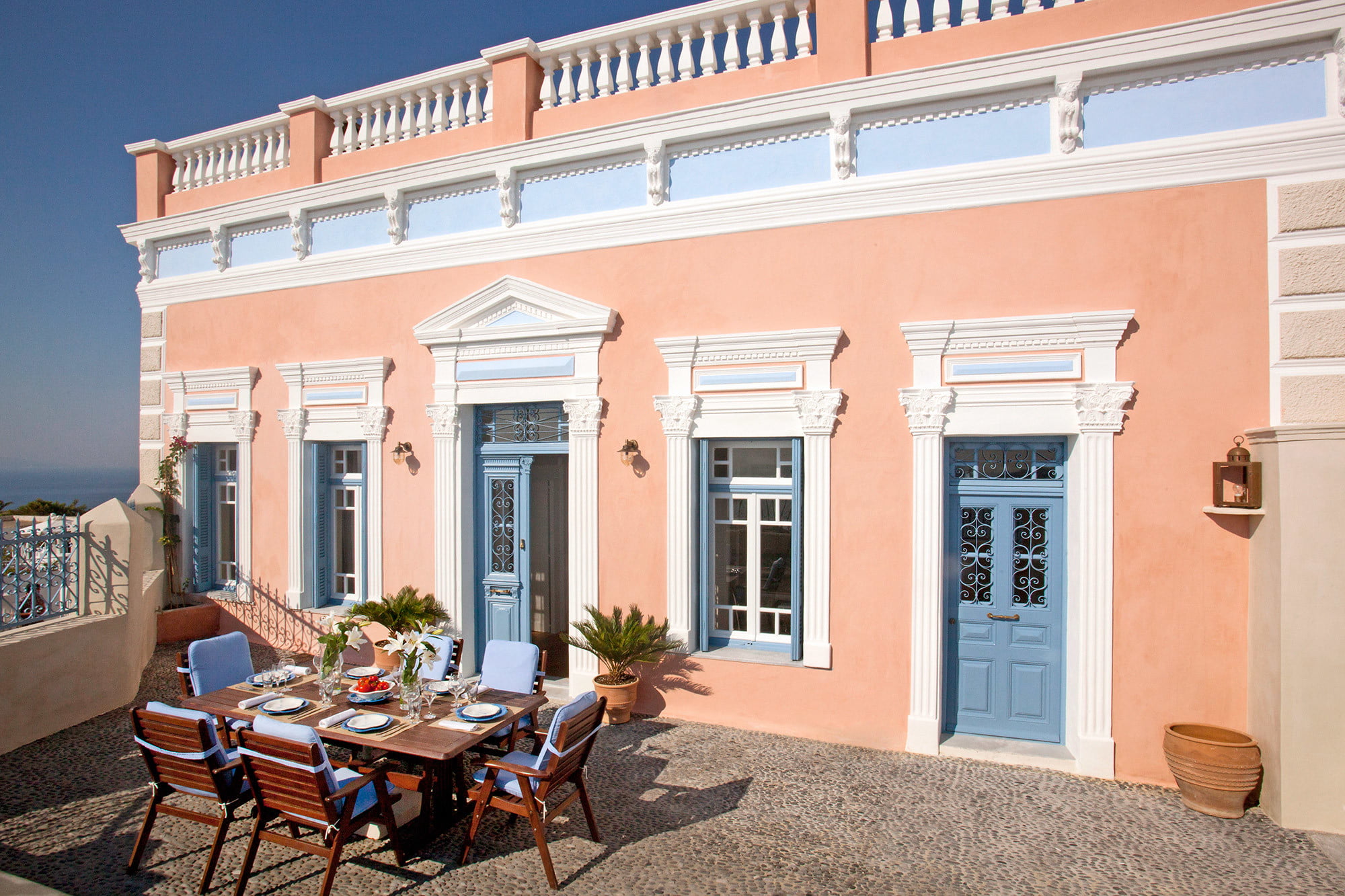 The neoclassical facade of the Capitan's Villa in Oia within a gated courtyard with an outdoor dining table.
