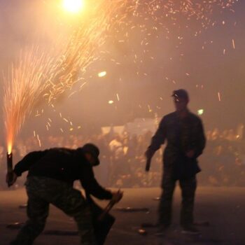 Kalamata Saitopolemos | Easter in Greece A man hold a custom rocket that is lit while he is kneeling and getting ready to light the other rocket held in his other hand. Close by is a second man getting ready to light his rockets. In the background the crowd has come to watch this unique Greek Easter local customs, part of celebrating a traditional Easter in Greece in the region of Kalamata.