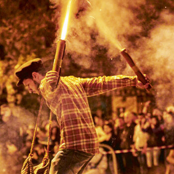 Kalamata Saitopolemos | Easter in Greece A young man wearing jeans and a checkered shirt is holding custom rockets that are lit in each hand. From his neck there is a traditional Greek bag hanging that contain even more custom made rockets. Crowds of people can be seen in the background that are there to celebrate Easter and watch this unique local Greek customs.