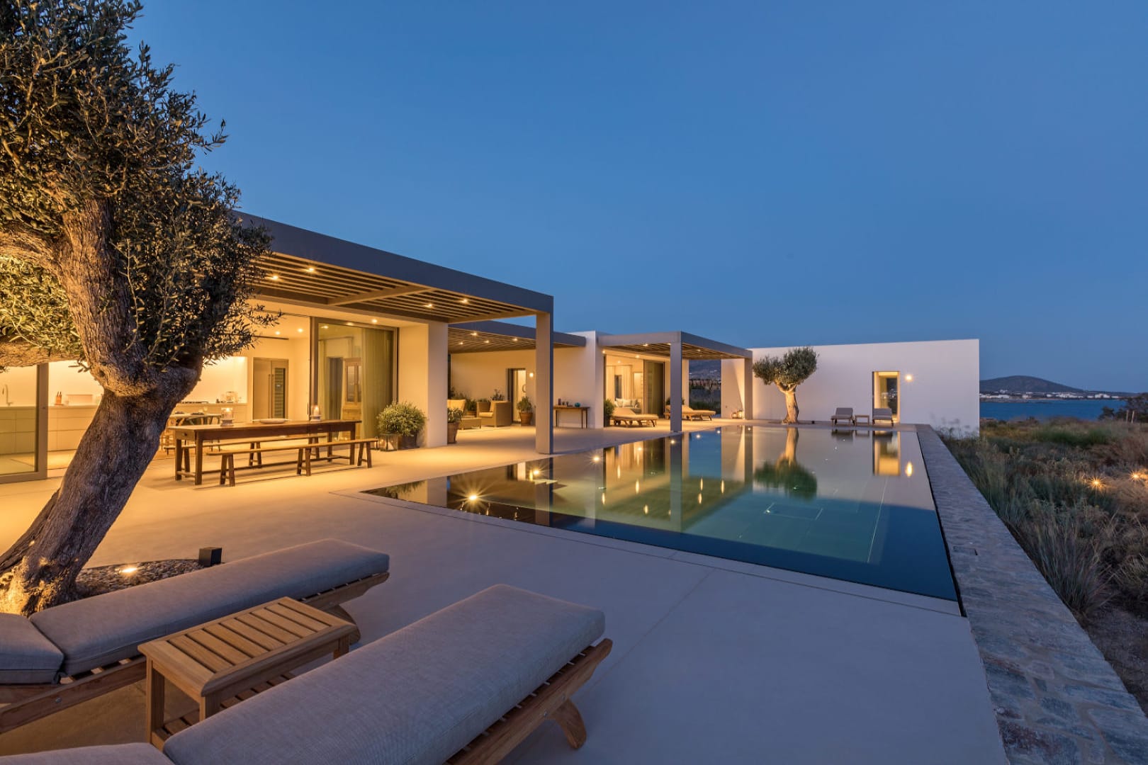 The facade of the exclusive Villa Olivia in Paros as seen from the black granite clad pool at dusk. behind the pool all lights are on emphasising the outdoor spaces filled with outdoor seating areas such as dining area, lounge area and sunbathing areas