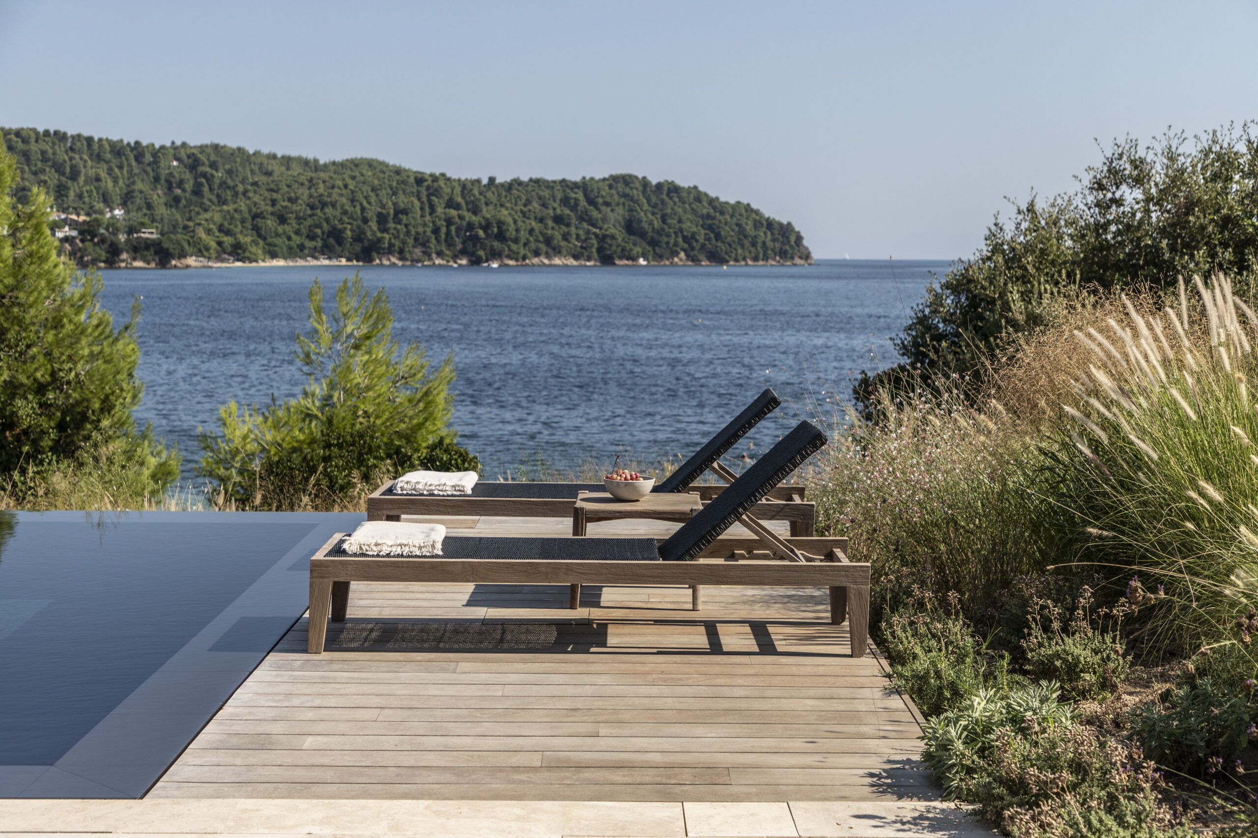 Two wooden sunbeds by a black granite clad pool found in one of the villas available for rent in the private villa collection by Rent A Greek Villa.