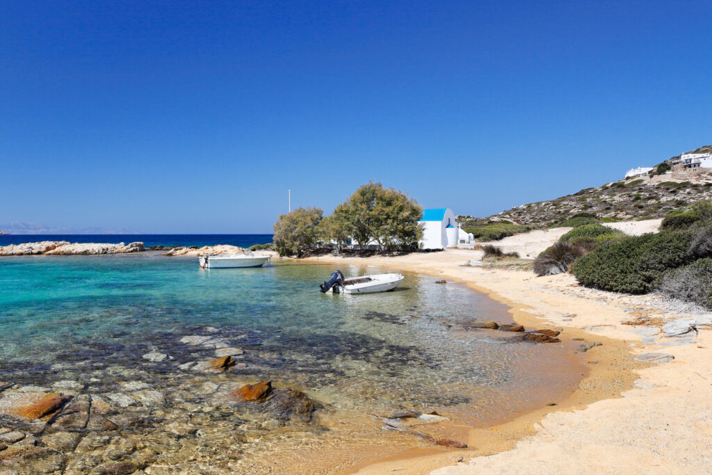 Antiparos guide | Top 10 Beaches by Rent A Greek Vilal A sandy share with a small boat in the cove, one of the top 10 beaches in Antiparos. In the background there is a small white church with a blue dome.