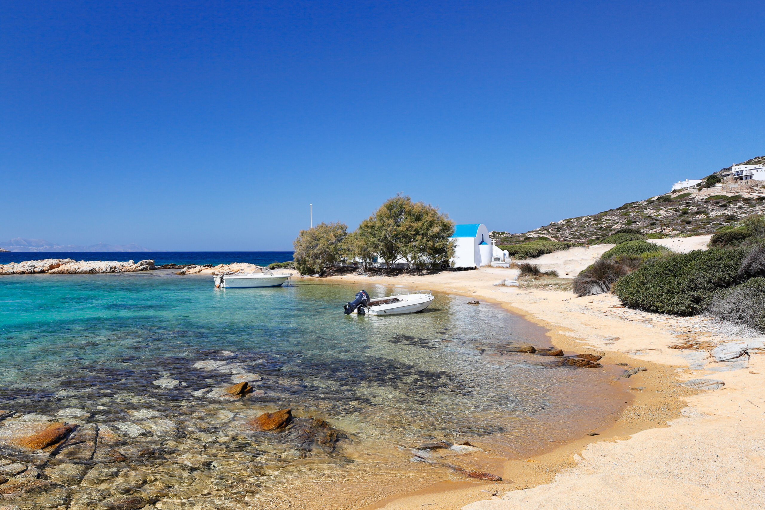 A sandy share with a small boat in the cove, one of the top 10 beaches in Antiparos. In the background there is a small white church with a blue dome.